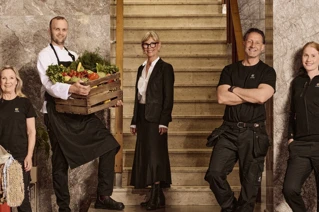 Team of service staff with diverse roles posing in stairwell, including a chef holding a crate of fresh vegetables, a housekeeper with cleaning supplies, a concierge in formal attire, and maintenance staff in uniforms.