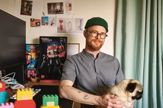 Smiling man with glasses and beanie holding a pug with LEGO toys and a Transformers collector's item in a cozy home office setup.