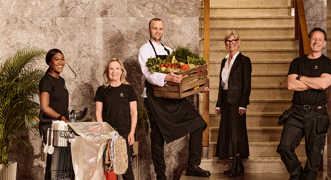 Diverse team of hotel staff posing with tools of their trades in a lobby with a marble wall and wooden stairs, including a housekeeper with cleaning equipment, a chef holding a crate of fresh vegetables, a manager, and a maintenance worker.