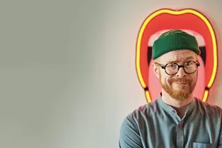Man with glasses and a green beanie smiling in front of a heart-shaped neon light.