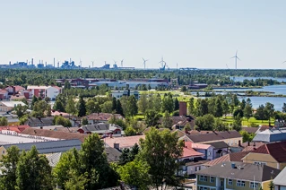 Panoramic view of a coastal town with red and yellow rooftops, wind turbines on horizon, and industrial skyline, depicting sustainable energy in urban landscapes.