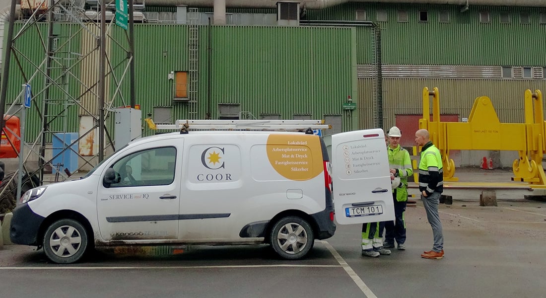 Två industriarbetare i varselvästar diskuterar vid en Coor servicebil på en industrianläggning.