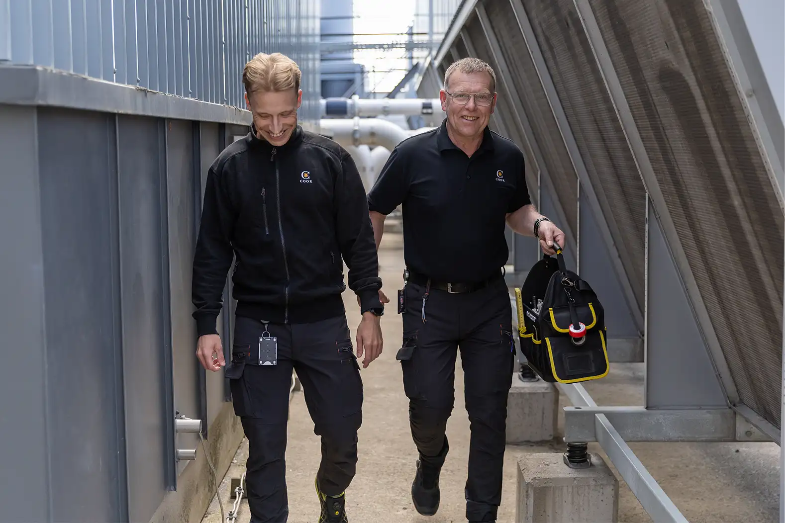 Två fastighetstekniker som går och ler i en industriell miljö
