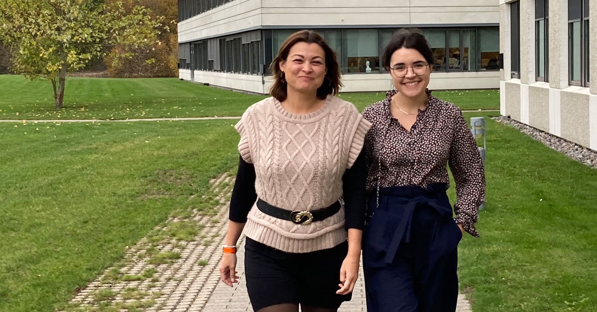 Two smiling women walking together outdoors near a modern building in autumn.