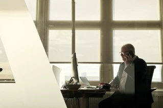 Man sitting in an office by a desk, talking on the phone.