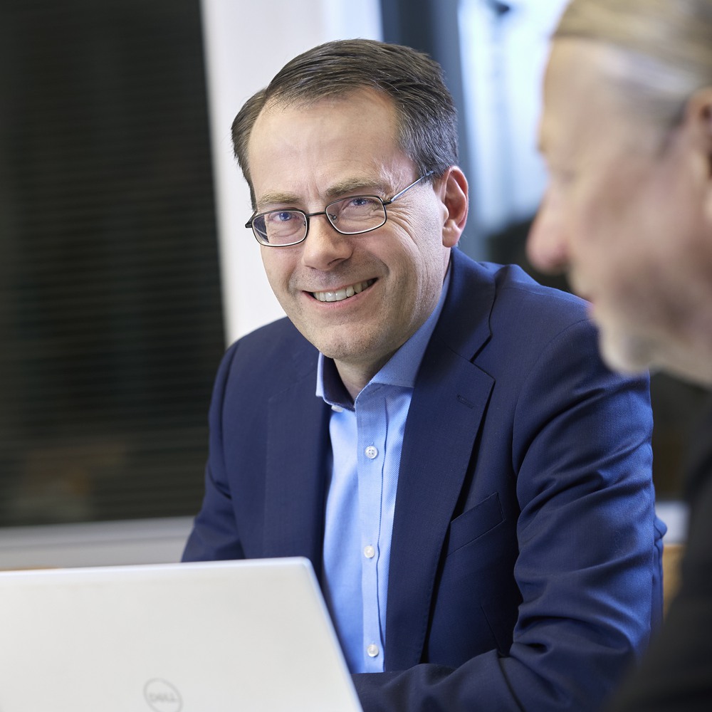 Professional man in glasses and blue suit smiling during a business meeting with colleague shown out of focus in foreground.