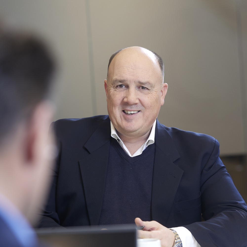 Smiling businessman in a suit having a conversation with a colleague out of focus in the foreground.