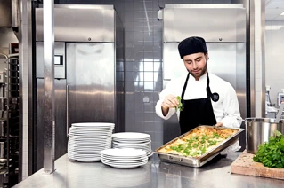 Male chef standing in a commercial kitchen putting fresh herbs on top of a gratine