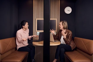 Two professional women engaging in a friendly conversation in a modern office booth with laptops and a minimalist clock on the wall.