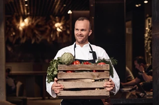 Chef holding a wooden crate full of fresh vegetables in a restaurant kitchen.