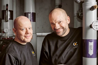 Two smiling technicians with black shirts featuring company logos, standing in an industrial setting with hydraulic equipment and pressure gauges.