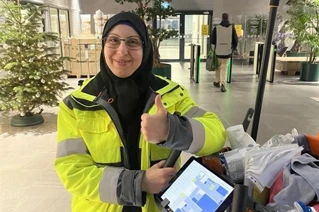 A woman in a yellow Coor-jacket giving a thumbs up next to a cleaning trolley.