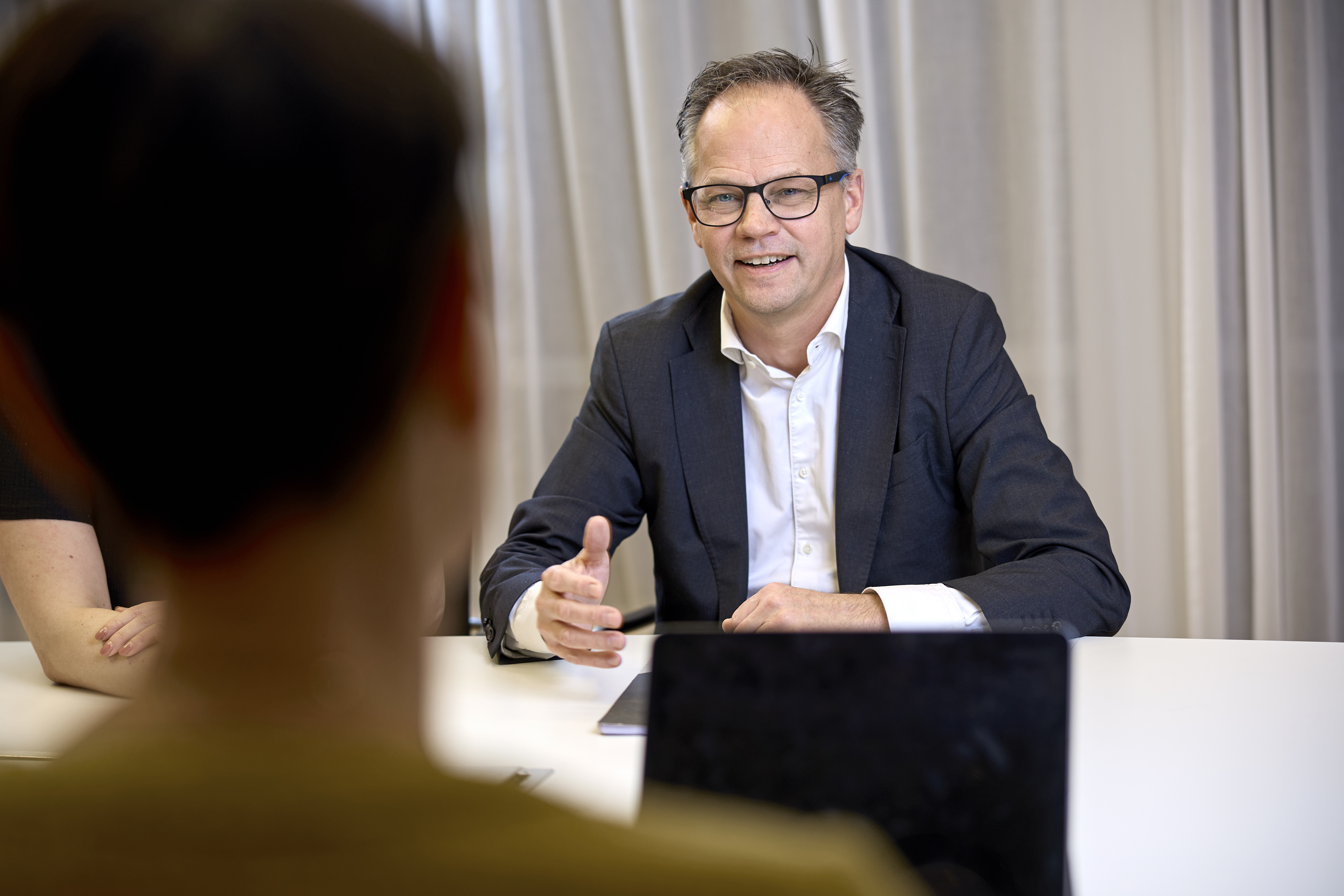 Professional middle-aged man with glasses smiling in a black suit and white shirt in conference room.