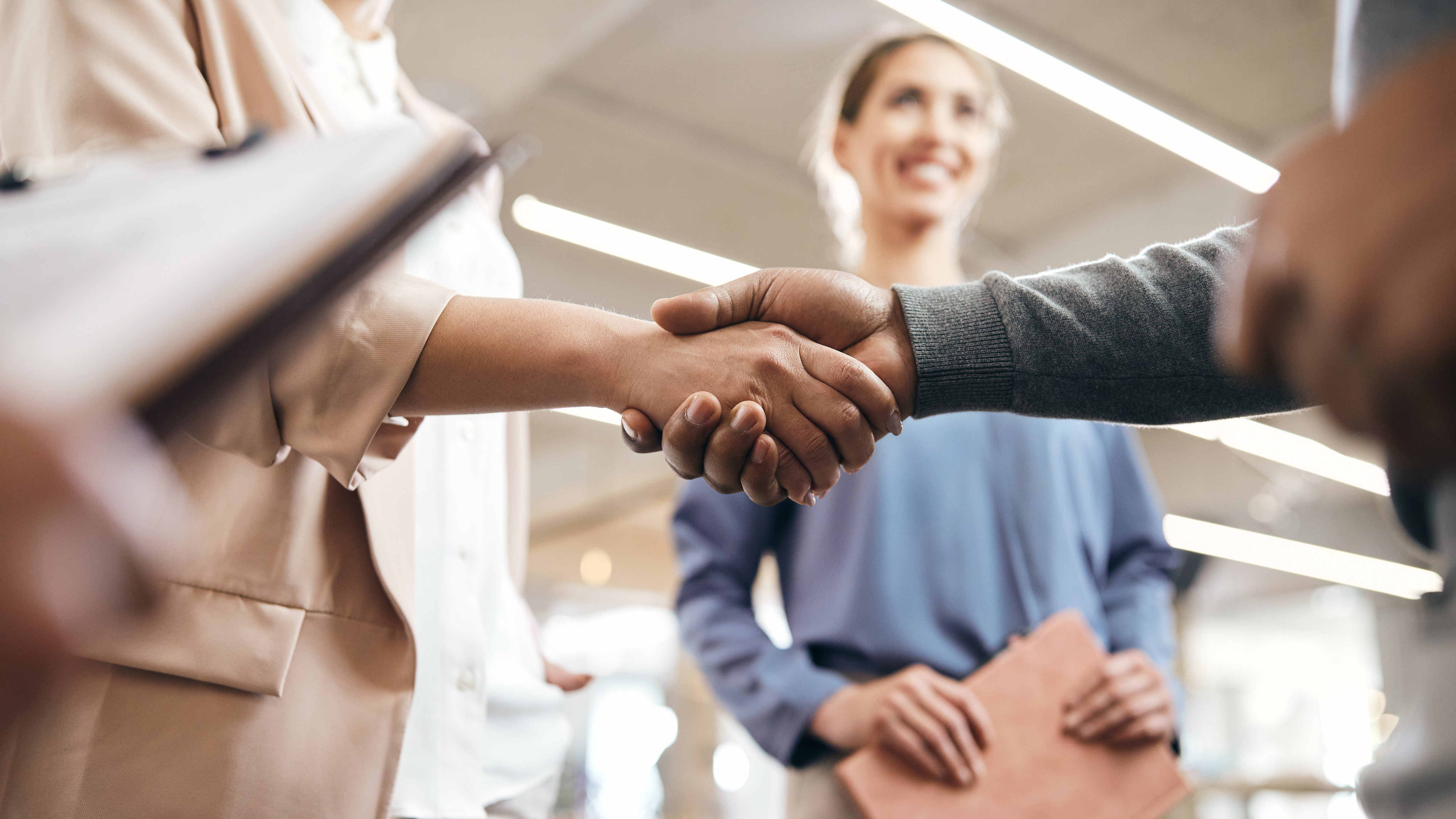 Two professionals shaking hands in a business setting with a smiling coworker in the background