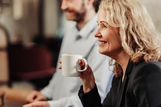 Woman in foreground holding a cup of hot beverage. A man can be seen in the background and it looks like he and the woman are part of a meeting.