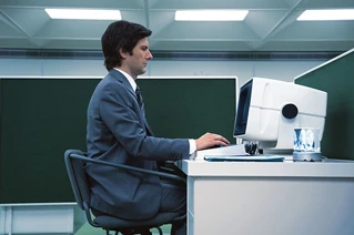 Man sitting by a desk, watching a computer screen, from the movie The Severance